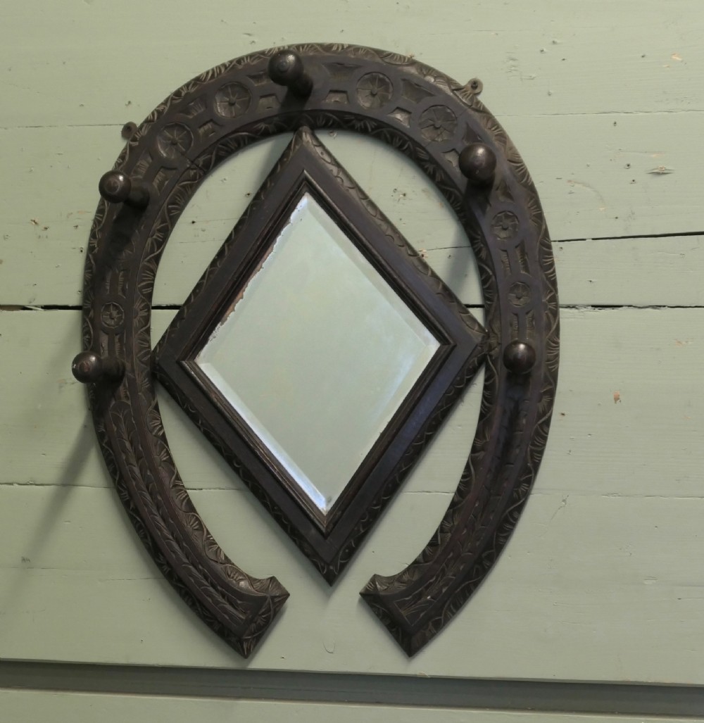 A dark carved oak horseshoe-shaped wall rack featuring five turned wooden coat pegs and a central diamond-shaped mirror.