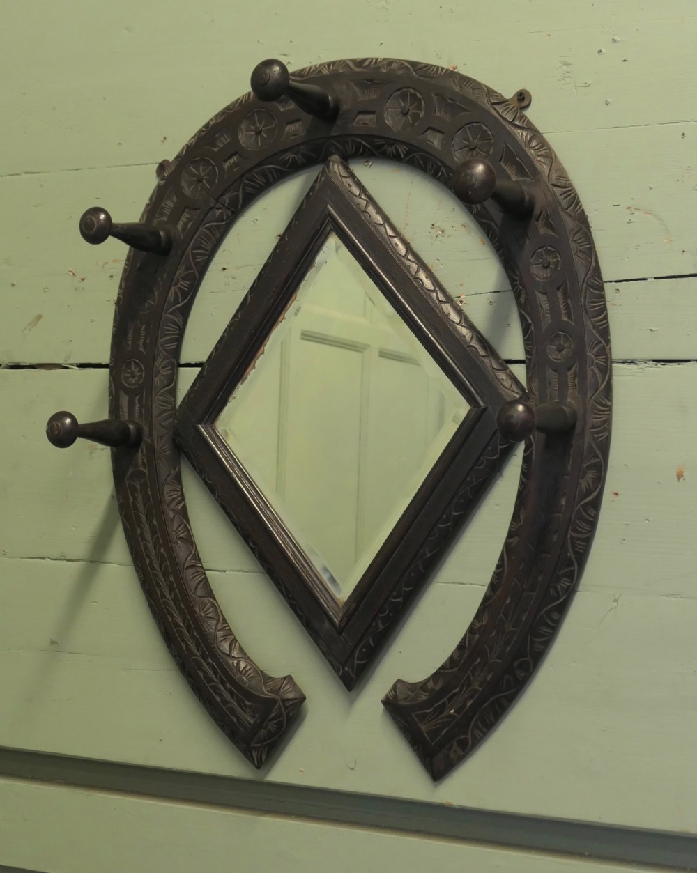A dark carved oak horseshoe-shaped wall rack featuring five turned wooden coat pegs and a central diamond-shaped mirror. View VI