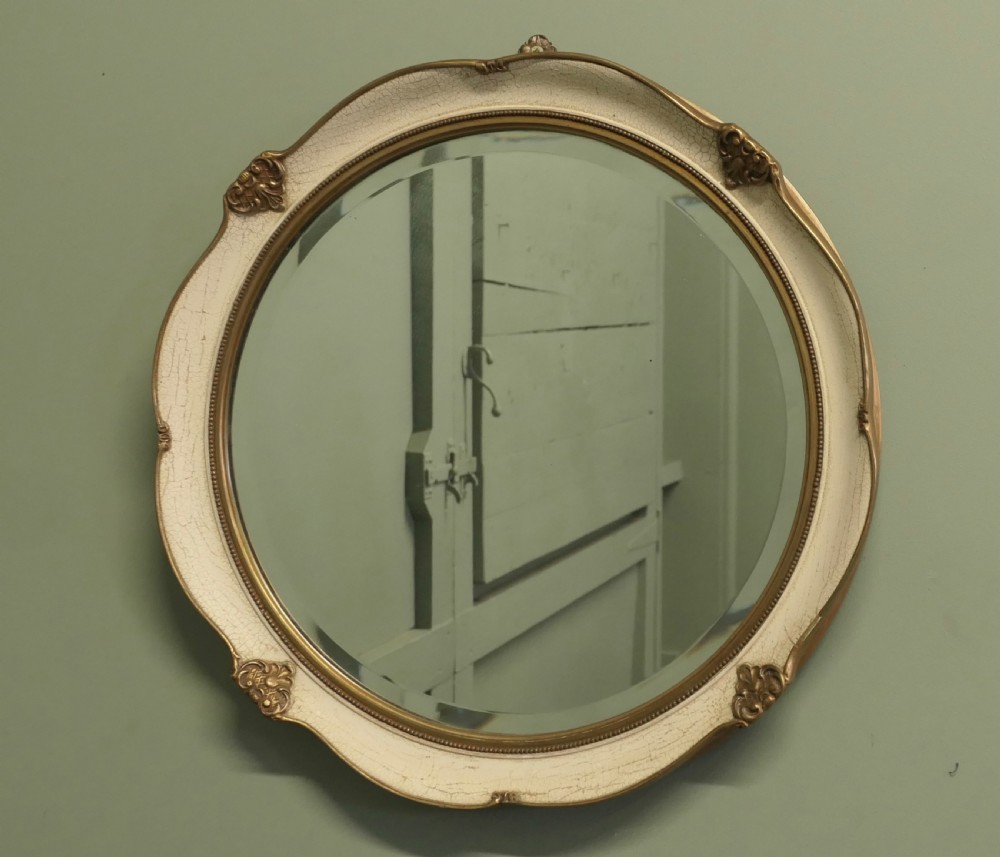 A circular wall mirror featuring a crackle-glazed cream painted frame accented with gilt embellishments and a bevelled glass plate.
