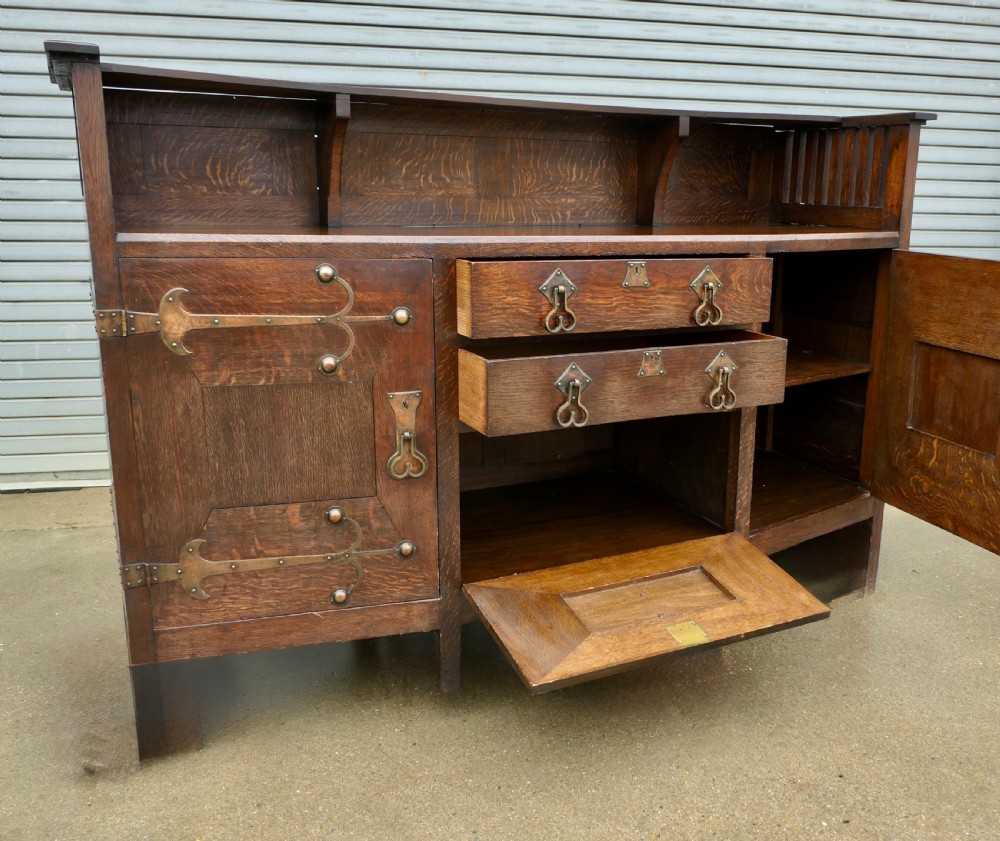 An Arts and Crafts oak sideboard featuring decorative copper strap hinges, heart-shaped drawer pulls, and a low shelf upper gallery. View IV