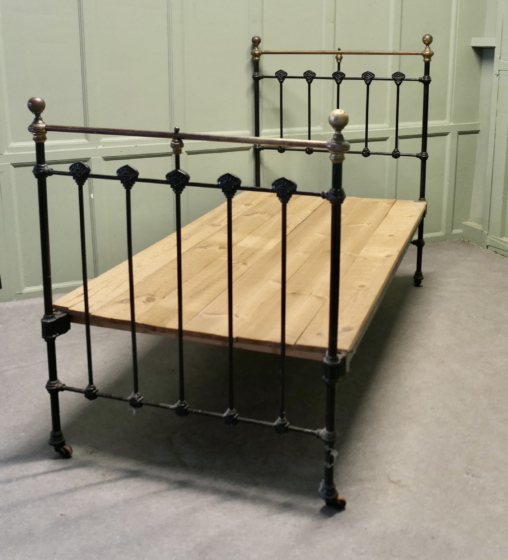 A Victorian single bedstead featuring a black painted iron frame with vertical spindles, brass top rails, brass finials, and a wooden slate base. View V