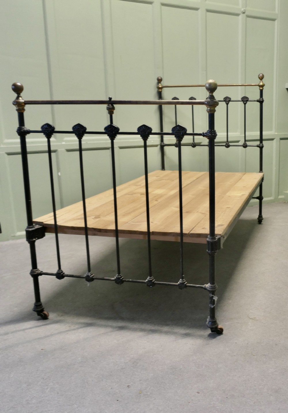 A Victorian single bedstead featuring a black painted iron frame with vertical spindles, brass top rails, brass finials, and a wooden slate base. View VI