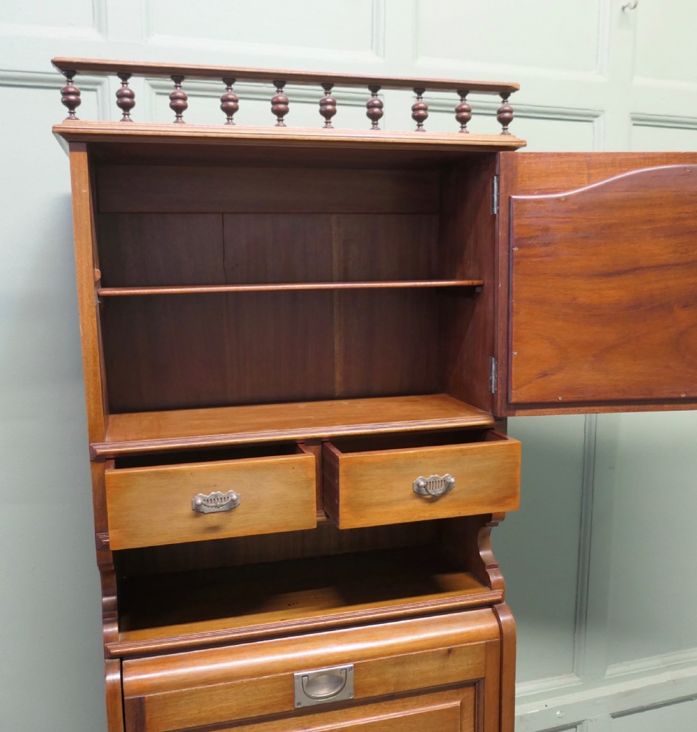 A tall, narrow wooden French marine vanity cabinet featuring a spindled top gallery, a mirrored upper cupboard, small drawers, a drop-front compartment, and lower storage doors. View II