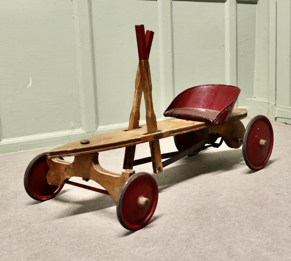 A 1930s hand-operated wooden toy vehicle with a pointed torpedo body, two red-tipped propulsion levers, a curved red seat, and four red wheels. View III