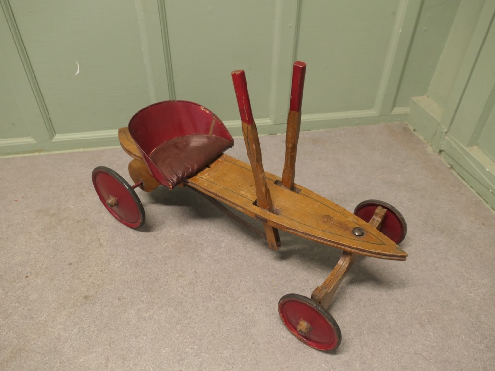A 1930s hand-operated wooden toy vehicle with a pointed torpedo body, two red-tipped propulsion levers, a curved red seat, and four red wheels. View IV