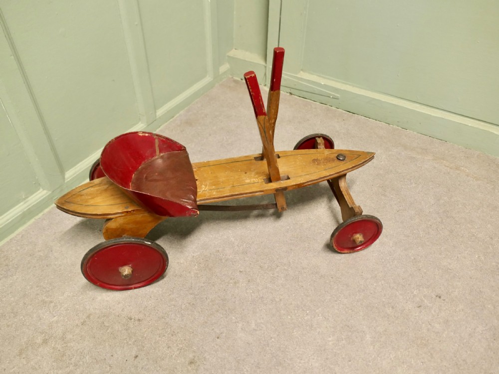 A 1930s hand-operated wooden toy vehicle with a pointed torpedo body, two red-tipped propulsion levers, a curved red seat, and four red wheels. View VI