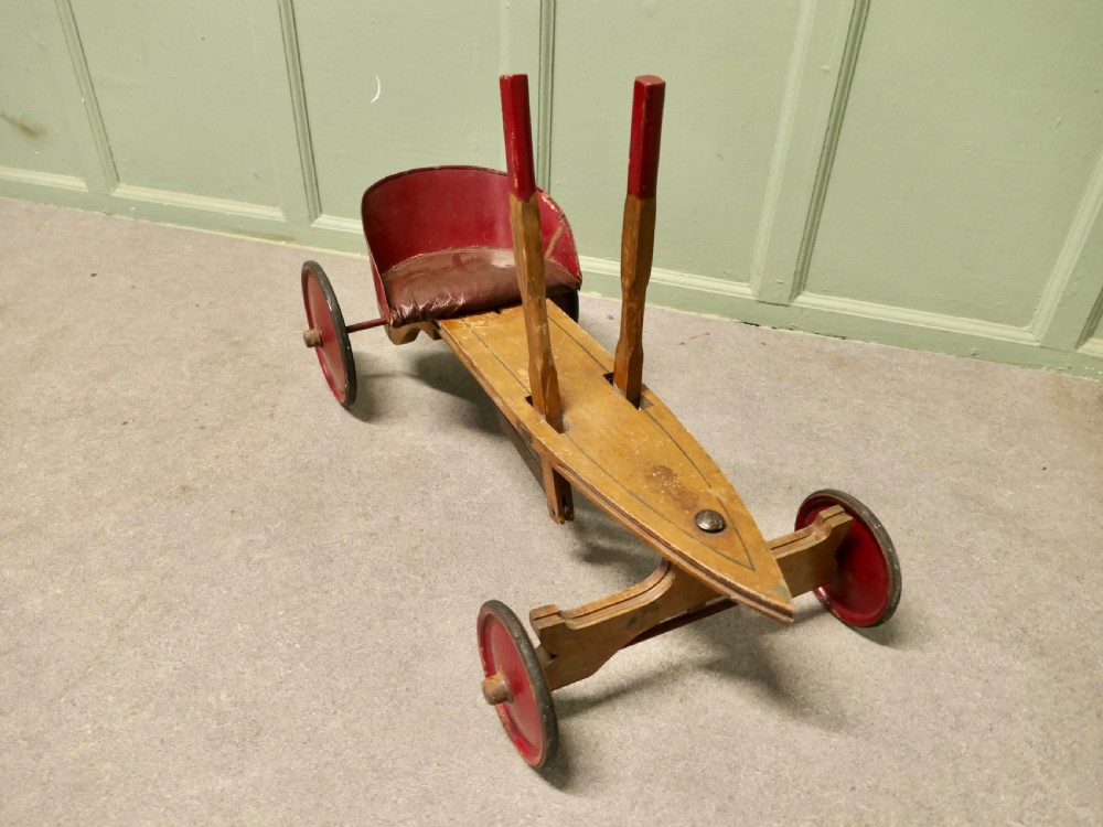A 1930s hand-operated wooden toy vehicle with a pointed torpedo body, two red-tipped propulsion levers, a curved red seat, and four red wheels. View VII