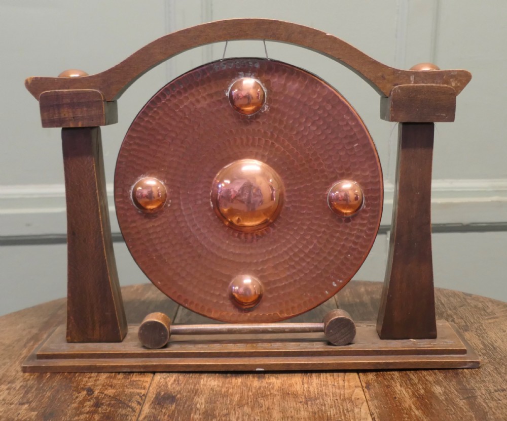 A circular hammered copper gong with raised bosses supported by an arched wooden frame and base with a small striker.