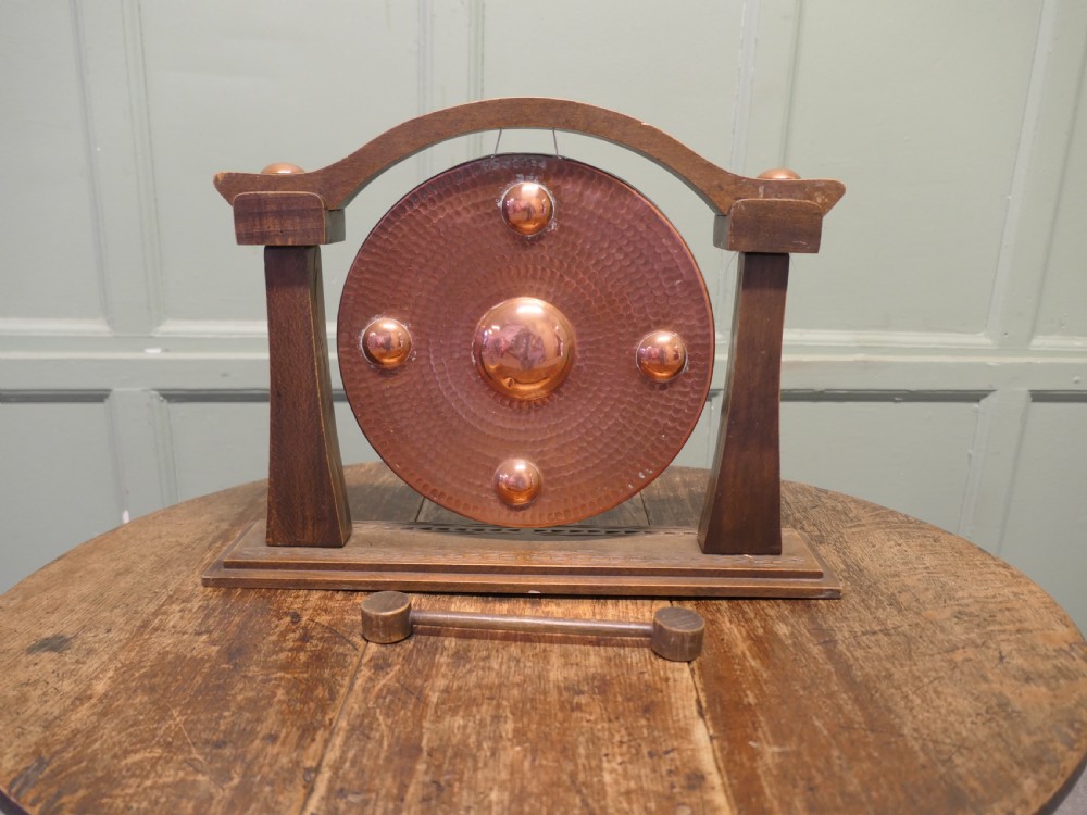 A circular hammered copper gong with raised bosses supported by an arched wooden frame and base with a small striker. View II