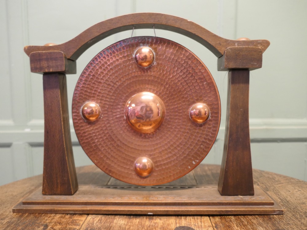 A circular hammered copper gong with raised bosses supported by an arched wooden frame and base with a small striker. View III