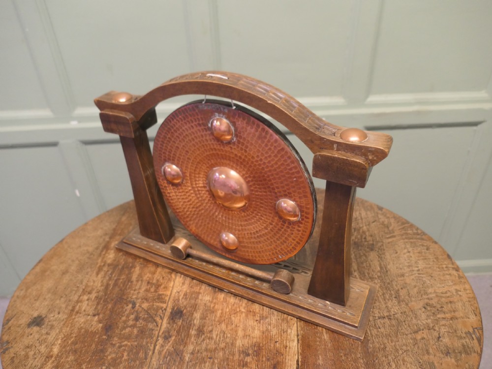 A circular hammered copper gong with raised bosses supported by an arched wooden frame and base with a small striker. View V