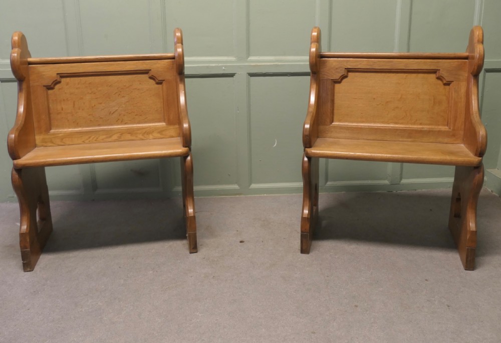 A pair of solid oak Arts and Crafts style hall chairs featuring shaped bench ends, fielded back panels, and gothic cut-outs.
