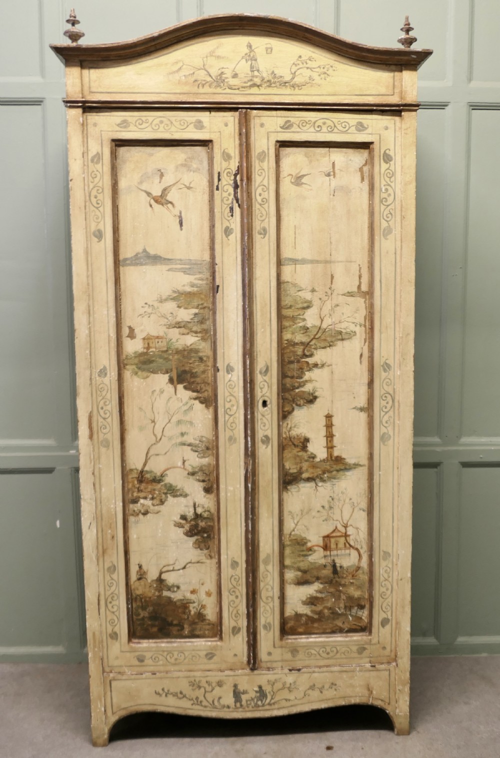 A tall, cream-painted antique cupboard featuring hand-painted Chinoiserie landscape scenes across two full-length doors and an arched pediment.
