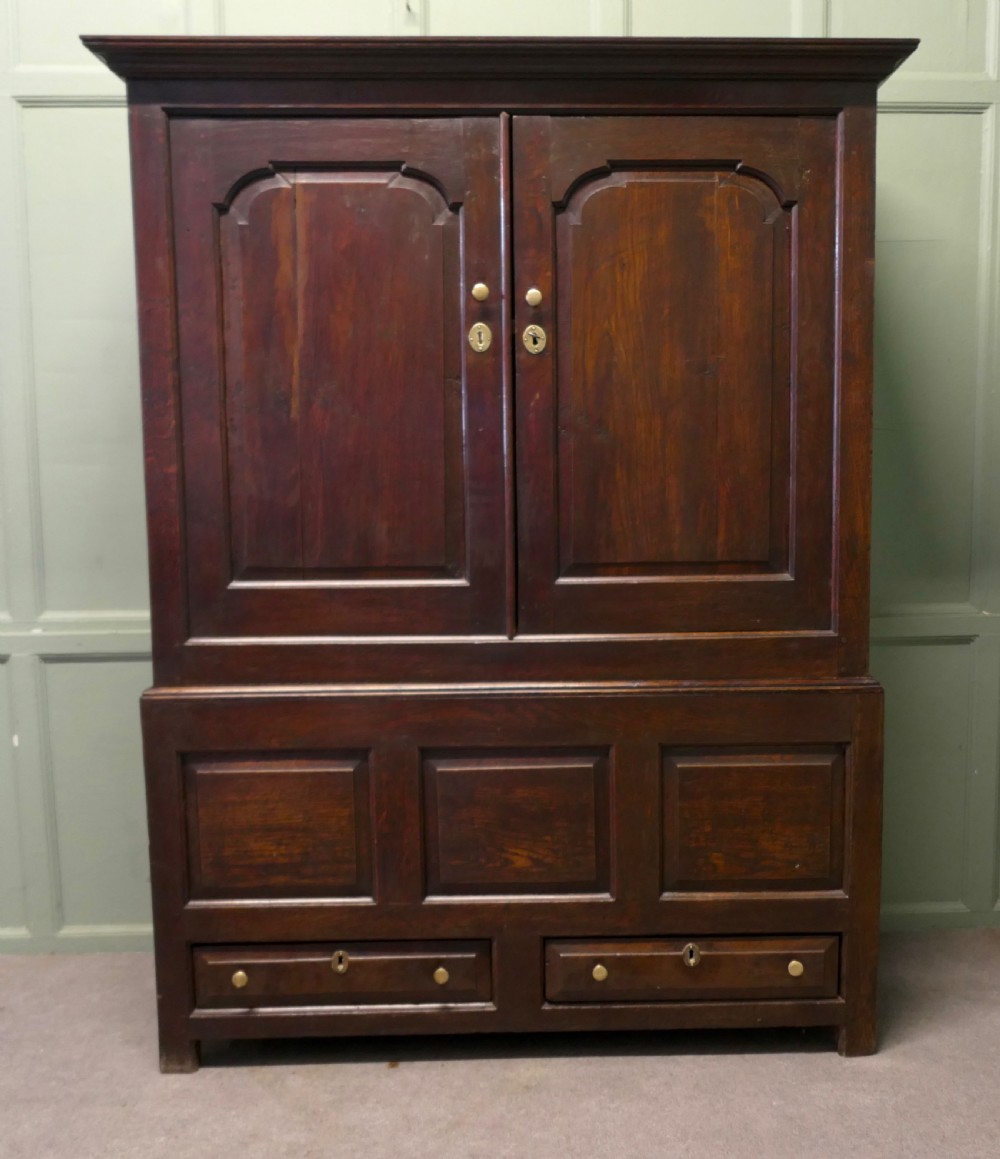 A tall dark Georgian oak housekeepers cupboard featuring two upper doors with arched fielded panels over a lower section with two drawers.