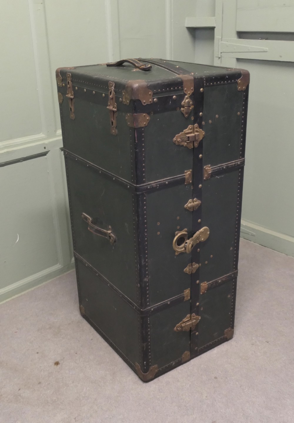 A tall, upright dark green French steamer wardrobe trunk with brass corner braces, studded trim, and side leather handles.
