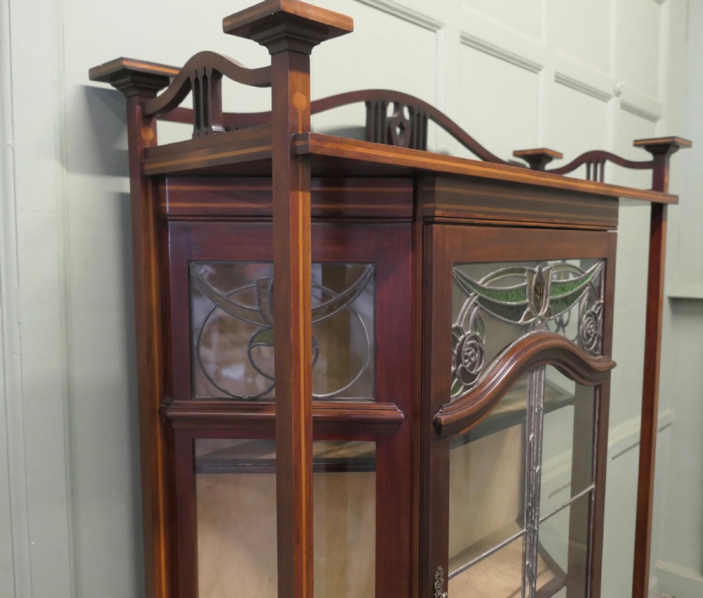 An Art Nouveau dark mahogany display cabinet featuring leaded stained glass panels with stylized floral motifs, inlaid wood detailing, and a gallery cresting. View II