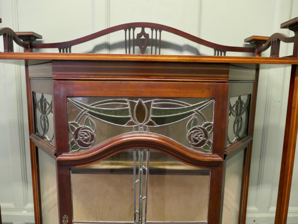 An Art Nouveau dark mahogany display cabinet featuring leaded stained glass panels with stylized floral motifs, inlaid wood detailing, and a gallery cresting. View IV