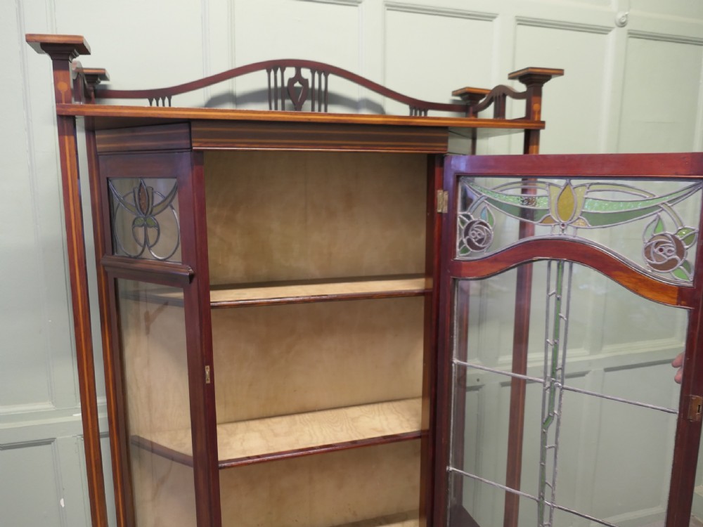 An Art Nouveau dark mahogany display cabinet featuring leaded stained glass panels with stylized floral motifs, inlaid wood detailing, and a gallery cresting. View V