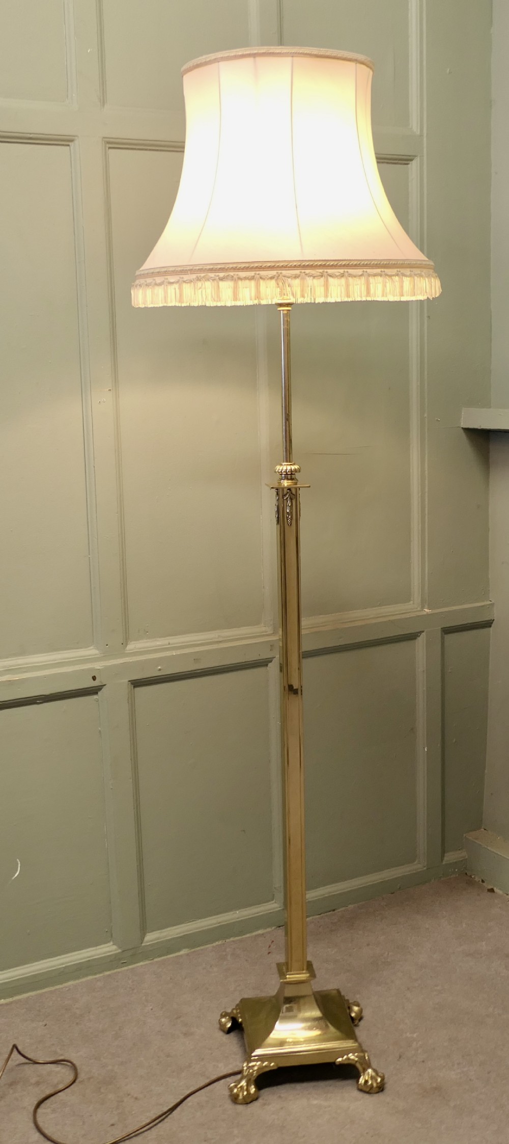 A tall brass telescopic floor lamp featuring a square column stem, stepped base with claw feet, and a cream fabric shade with fringe.