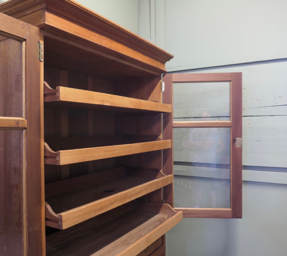An Edwardian warm pine linen press with a glazed two-door upper cupboard revealing internal shelves above a base chest of four drawers. View IV