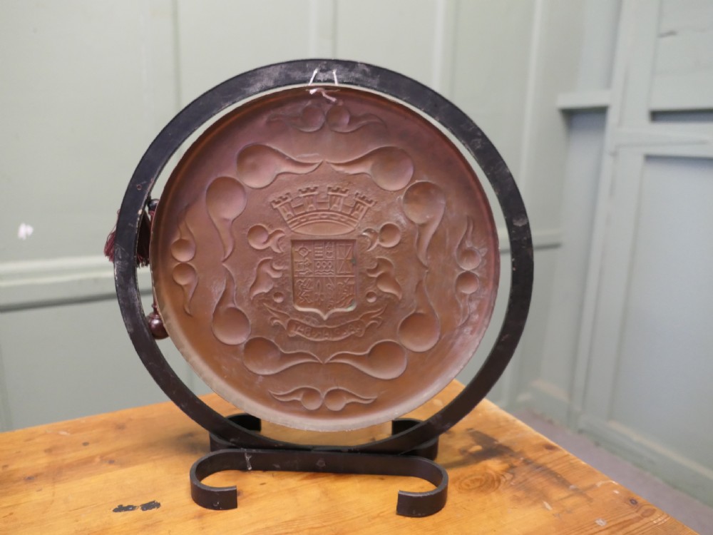 A circular hand-hammered copper table gong featuring a Basque heraldic crest motif, supported inside a dark textured iron circular stand with an attached striker. View II