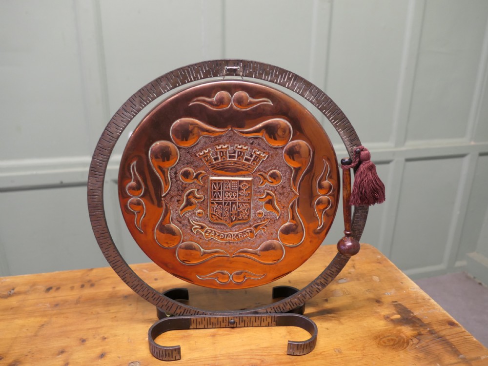 A circular hand-hammered copper table gong featuring a Basque heraldic crest motif, supported inside a dark textured iron circular stand with an attached striker. View III