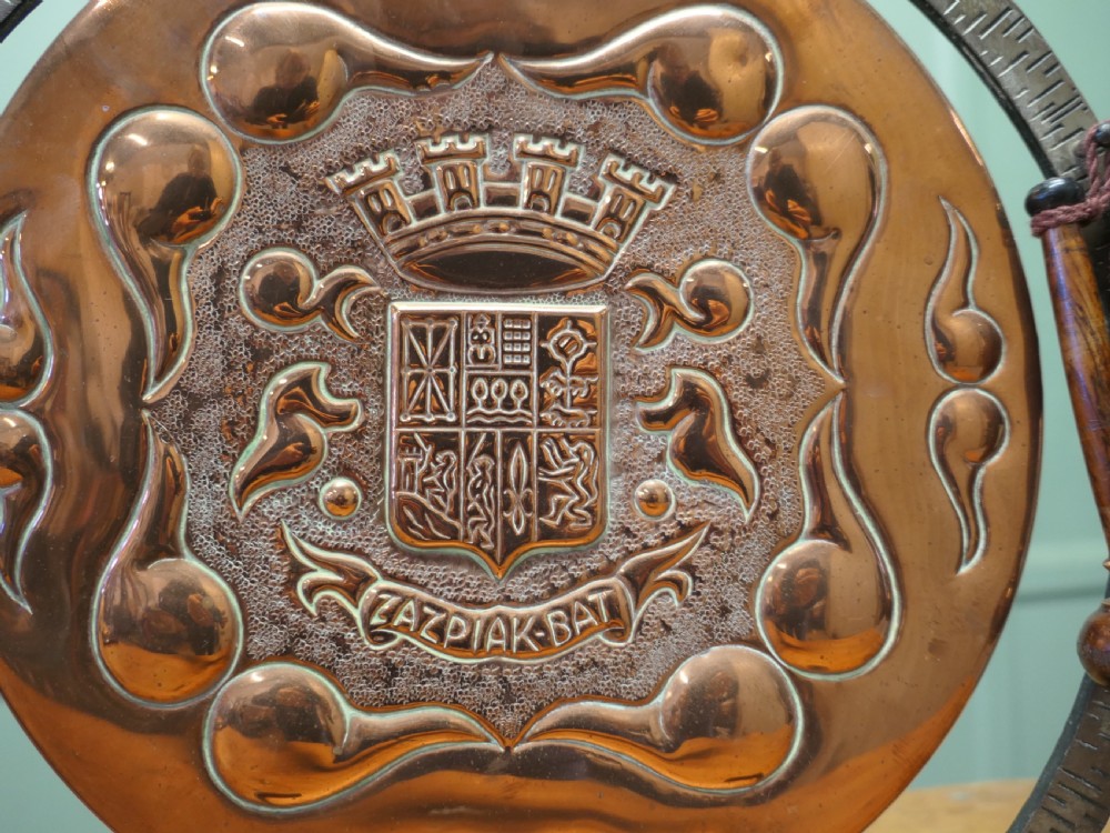 A circular hand-hammered copper table gong featuring a Basque heraldic crest motif, supported inside a dark textured iron circular stand with an attached striker. View IV