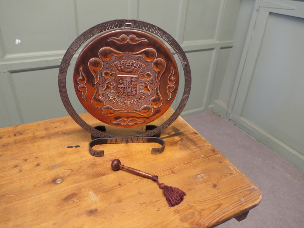 A circular hand-hammered copper table gong featuring a Basque heraldic crest motif, supported inside a dark textured iron circular stand with an attached striker. View V