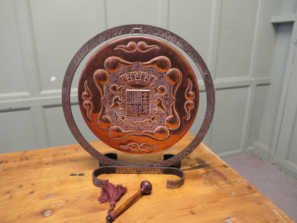 A circular hand-hammered copper table gong featuring a Basque heraldic crest motif, supported inside a dark textured iron circular stand with an attached striker. View VI