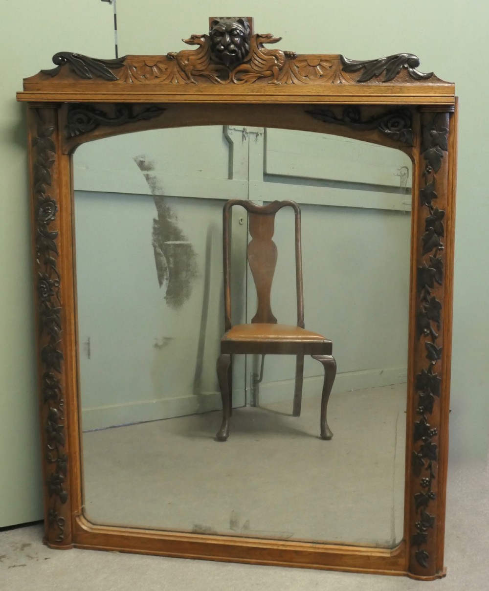 A large rectangular oak wall mirror featuring an arched glass top, relief foliate vine side carvings, and a crest with a carved lion mask.
