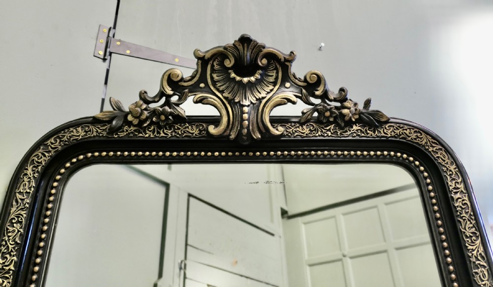 A tall rectangular ebonised pier mirror with rounded upper corners, an engraved gold leaf border, beaded moulding, and a carved central crest. View V