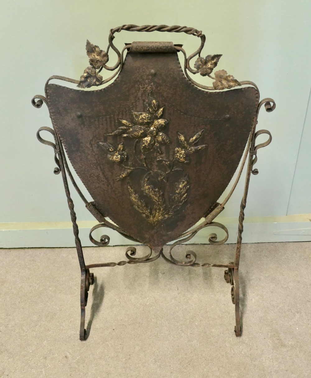 A wrought iron fire screen with a central shield-shaped metal panel featuring embossed gold-toned floral motifs and scrollwork decoration. View V