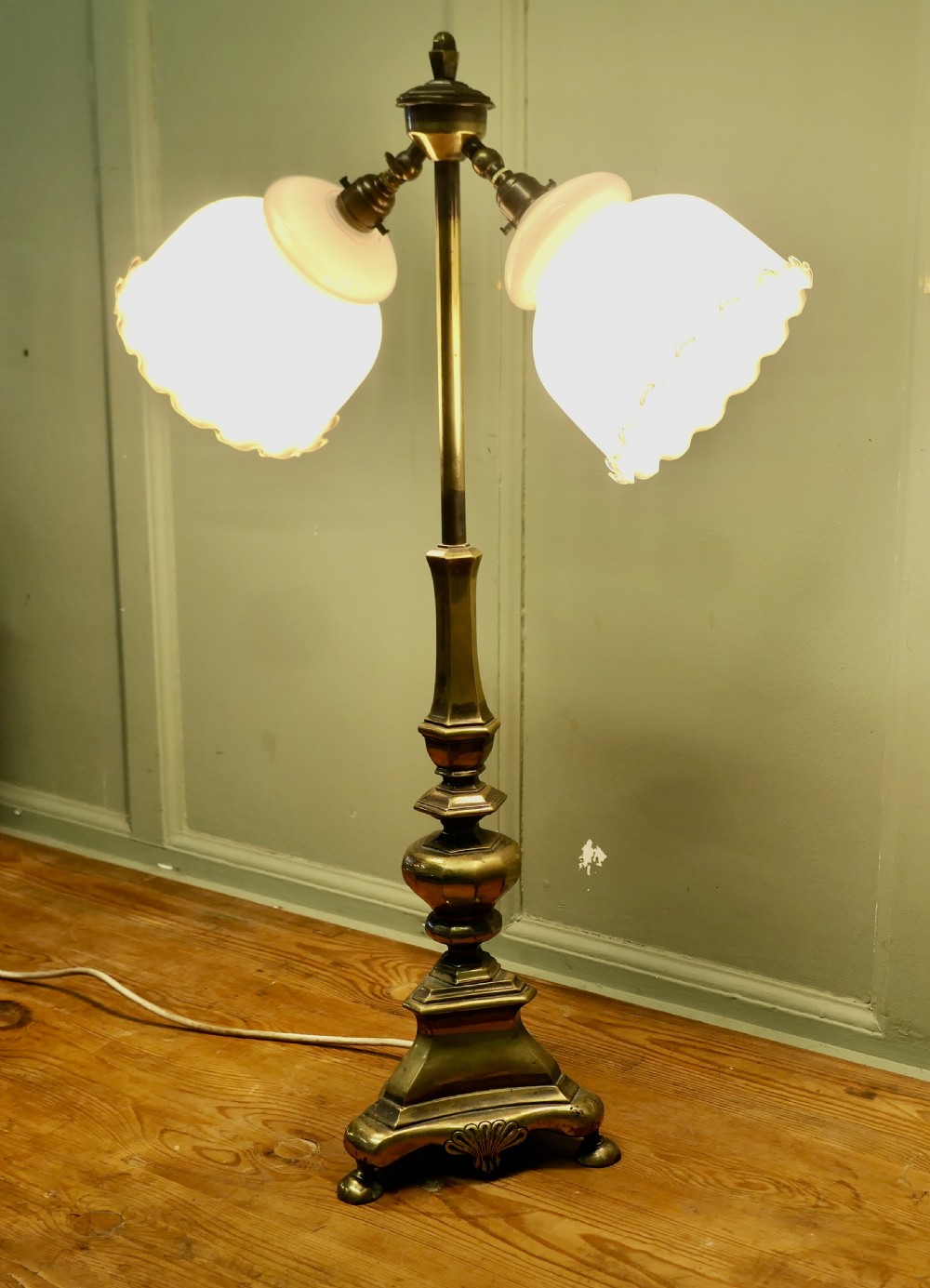 An antique brass table lamp with a stepped pedestal base, central turned column, and two angled arms supporting white frilled glass shades. View VI