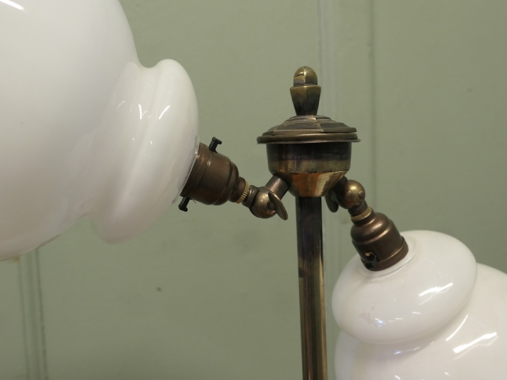 An antique brass table lamp with a stepped pedestal base, central turned column, and two angled arms supporting white frilled glass shades. View VII