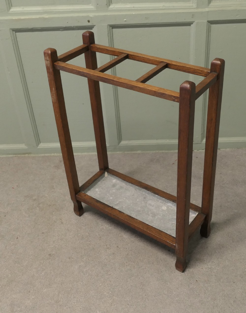 An Arts and Crafts golden oak stick stand featuring a rectangular frame with three divided top sections and a metal drip tray at the base.