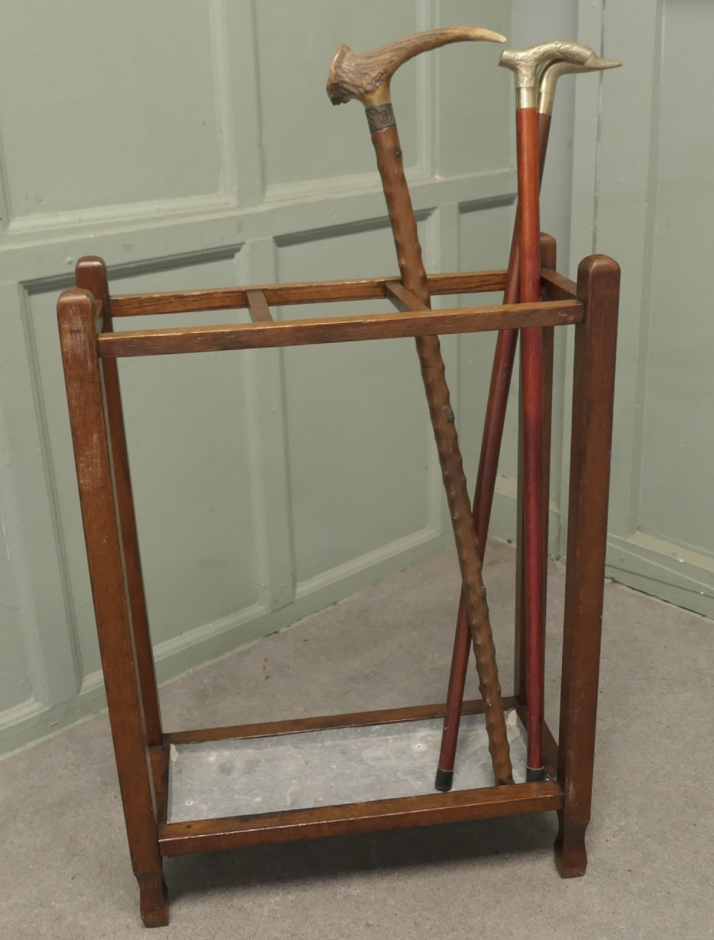 An Arts and Crafts golden oak stick stand featuring a rectangular frame with three divided top sections and a metal drip tray at the base. View III