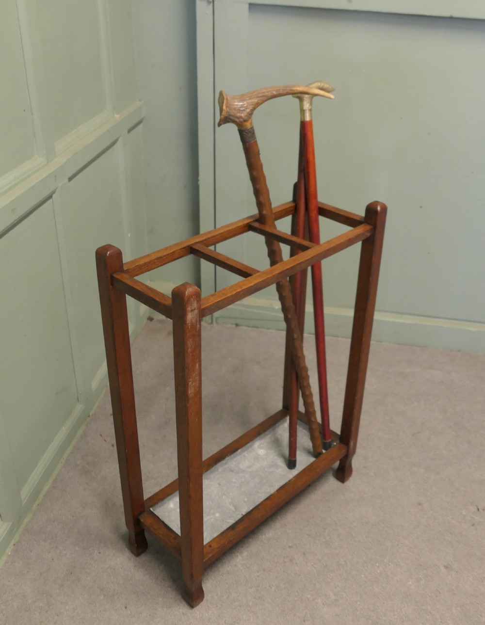 An Arts and Crafts golden oak stick stand featuring a rectangular frame with three divided top sections and a metal drip tray at the base. View IV