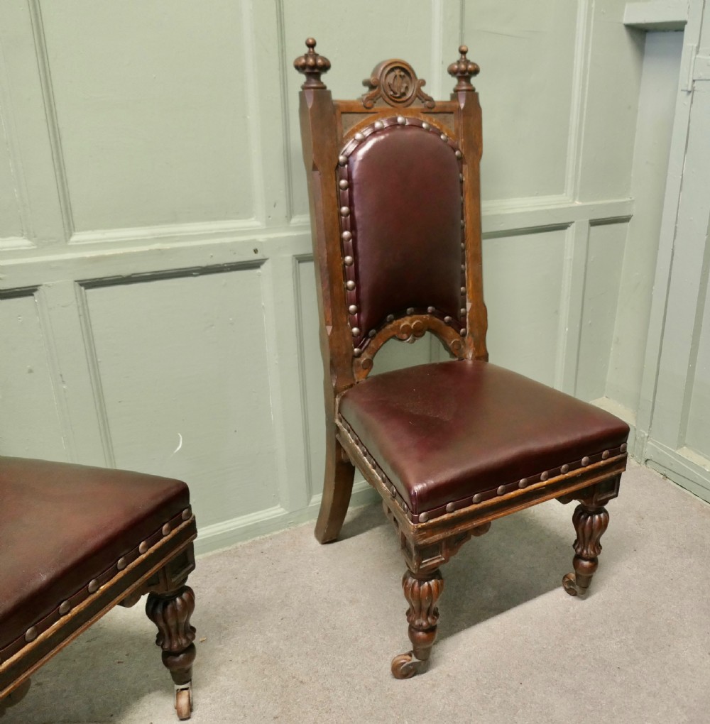 A set of six carved dark oak Gothic revival dining chairs featuring high upholstered leather backs, brass studs, and turned legs with castors. View II
