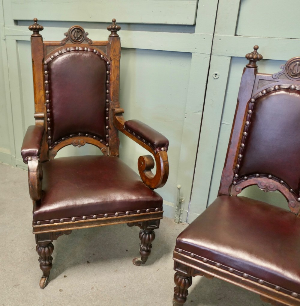 A set of six carved dark oak Gothic revival dining chairs featuring high upholstered leather backs, brass studs, and turned legs with castors. View V