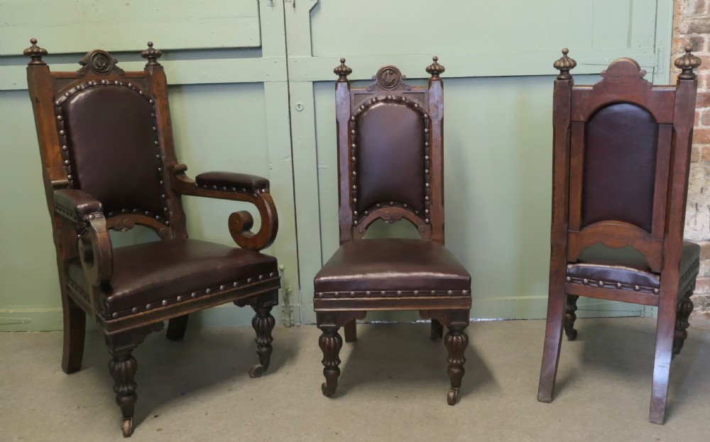 A set of six carved dark oak Gothic revival dining chairs featuring high upholstered leather backs, brass studs, and turned legs with castors. View VII
