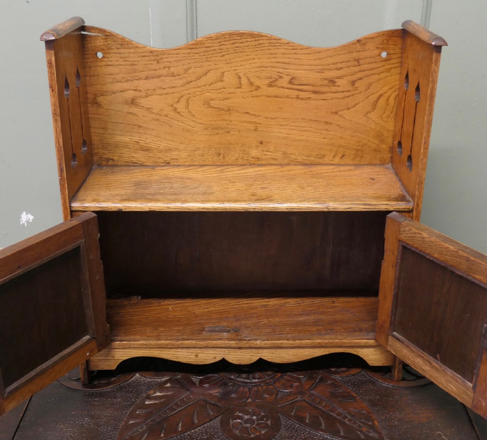An Arts and Crafts oak wall cabinet featuring twin panelled doors, a curved back gallery, and pierced spade motifs on the side panels. View II