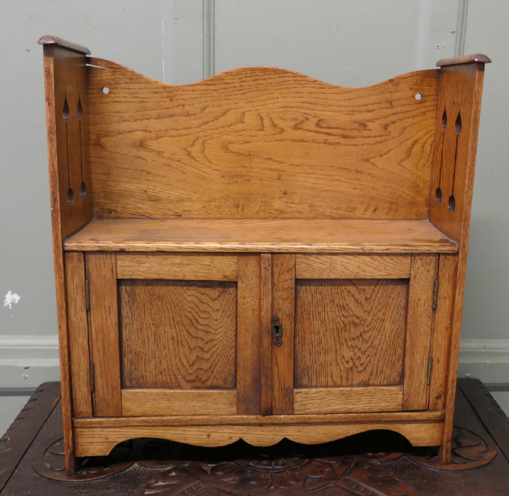 An Arts and Crafts oak wall cabinet featuring twin panelled doors, a curved back gallery, and pierced spade motifs on the side panels. View V