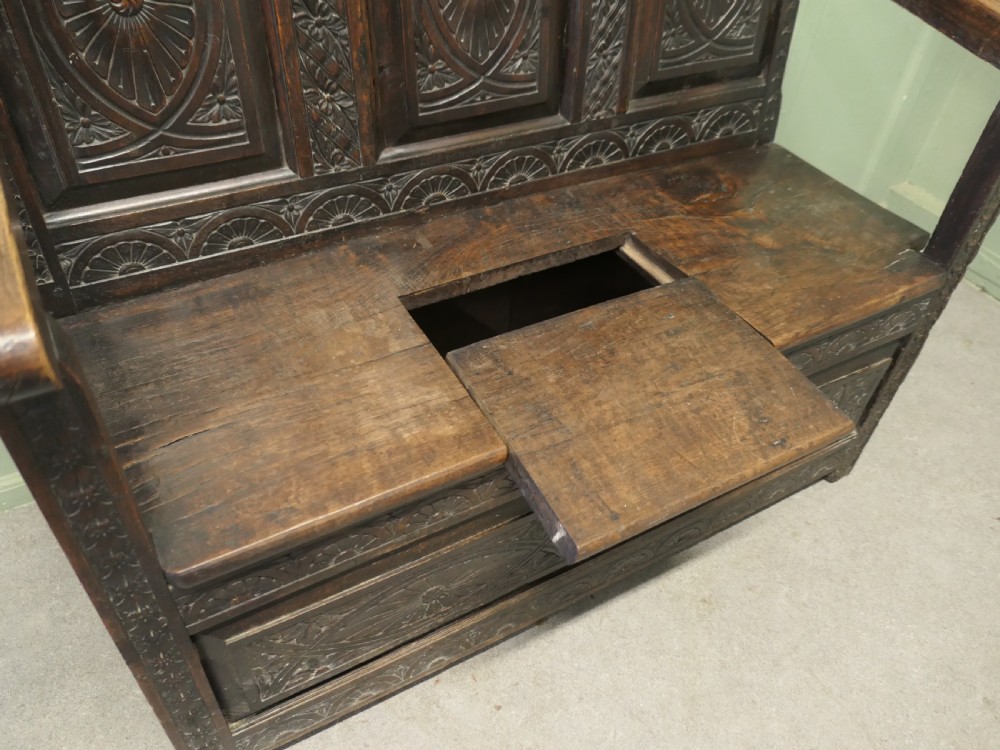 An eighteenth-century dark oak high back settle featuring intricately carved panels, straight armrests, and a lifted seat storage box. View II