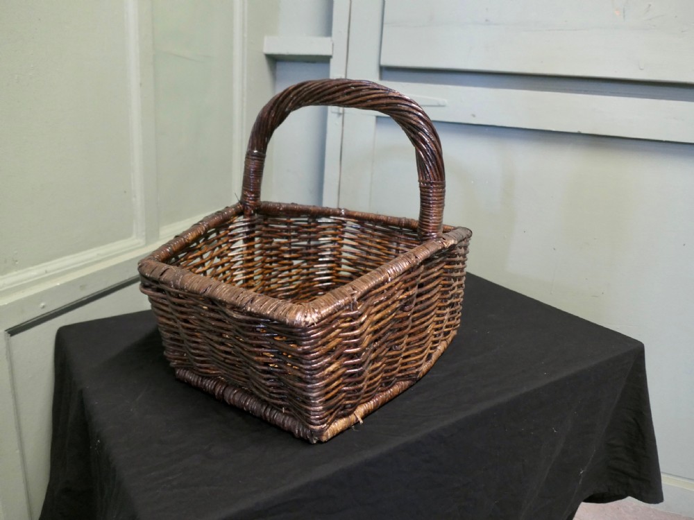 A dark brown woven wicker basket featuring a square base, flared sides, and a high twisted arched handle.