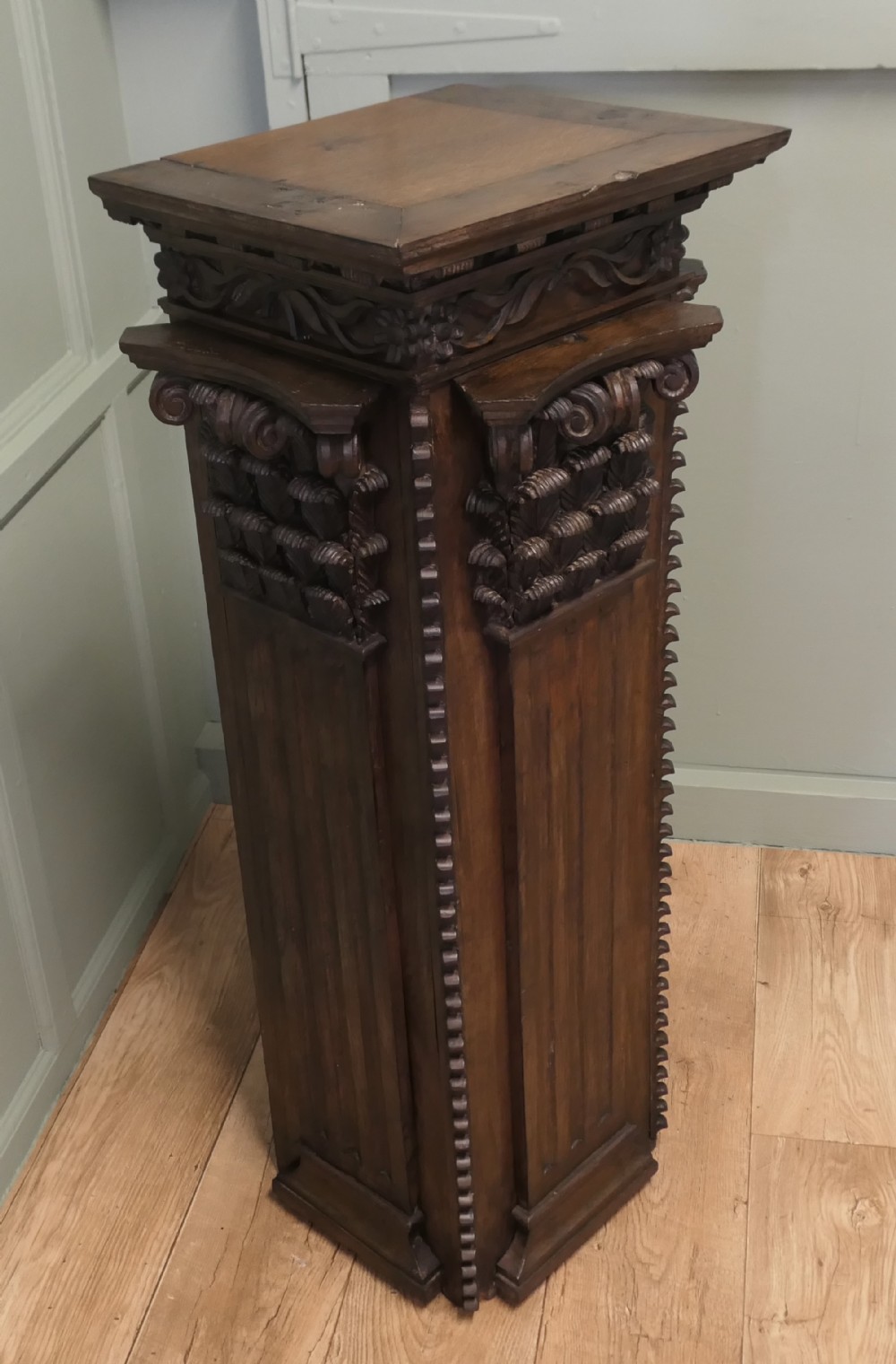 A dark carved oak column display pedestal featuring acanthus leaf detailing, volutes, and a square top surface.