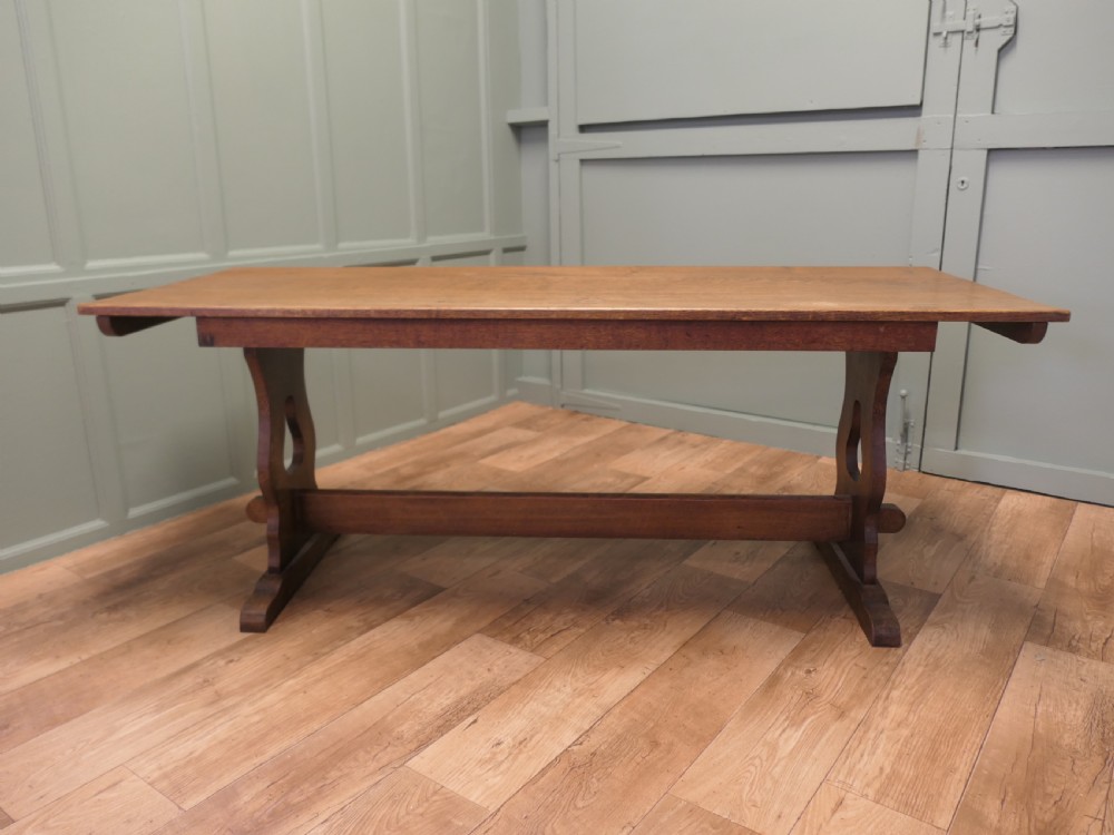 An Arts and Crafts solid golden oak refectory dining table featuring shaped trestle end supports and a central connecting stretcher.