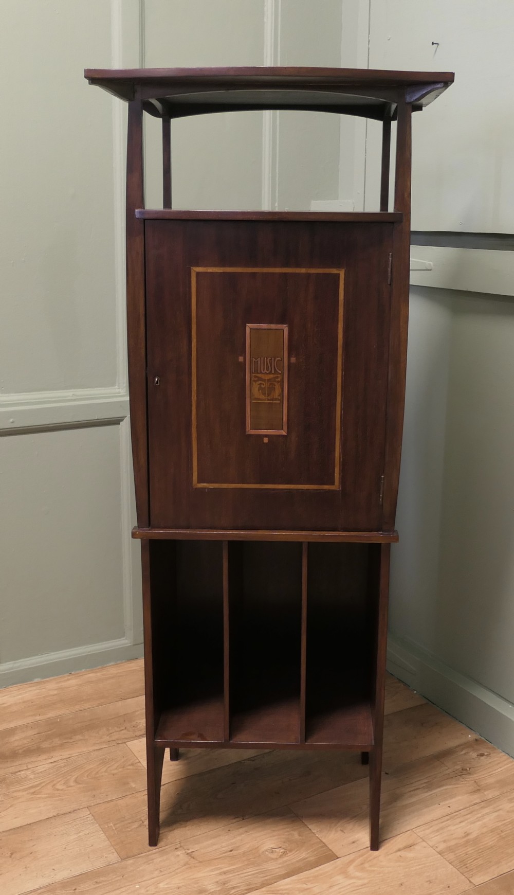 A tall dark wood Arts and Crafts music cabinet featuring an open top shelf, a central cupboard door with inlay reading MUSIC, and three open vertical slots below.