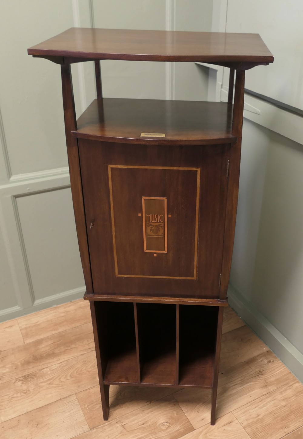A tall dark wood Arts and Crafts music cabinet featuring an open top shelf, a central cupboard door with inlay reading MUSIC, and three open vertical slots below. View V