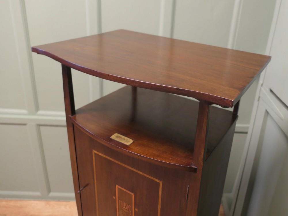 A tall dark wood Arts and Crafts music cabinet featuring an open top shelf, a central cupboard door with inlay reading MUSIC, and three open vertical slots below. View VI