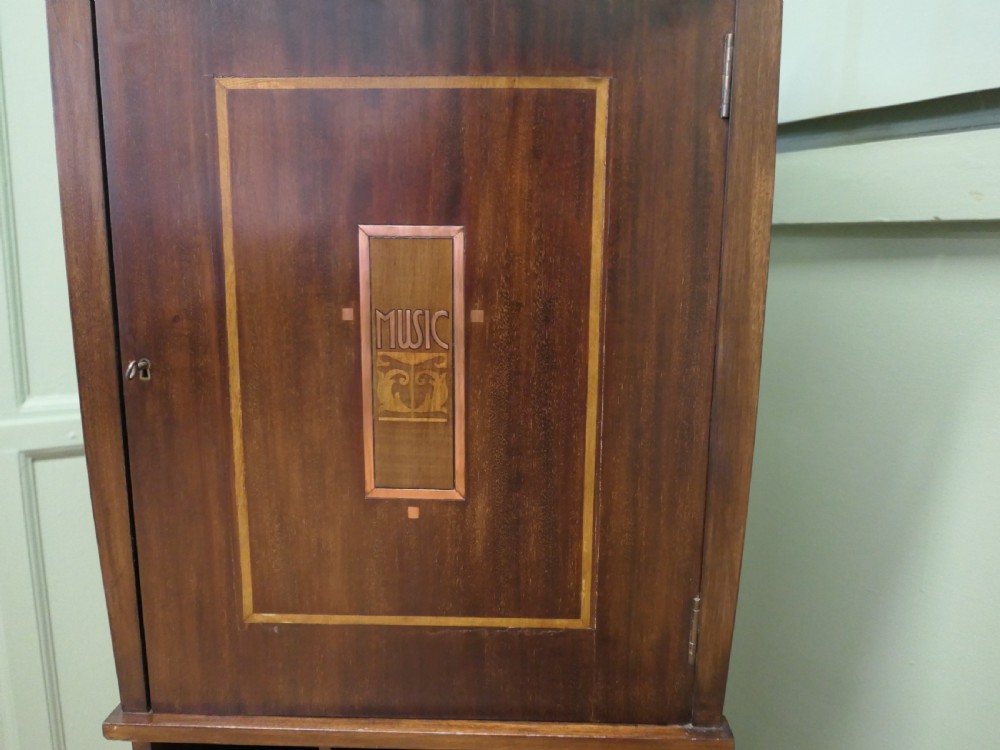 A tall dark wood Arts and Crafts music cabinet featuring an open top shelf, a central cupboard door with inlay reading MUSIC, and three open vertical slots below. View VII
